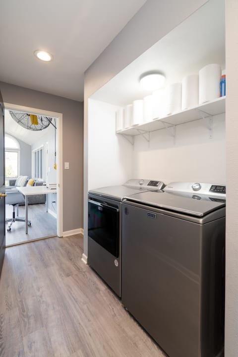 Modern laundry room with washer and dryer, paper towel storage, and a view of a bright sitting area.