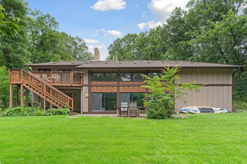 A two-story house surrounded by trees and a green lawn, featuring a staircase and a paddleboard.