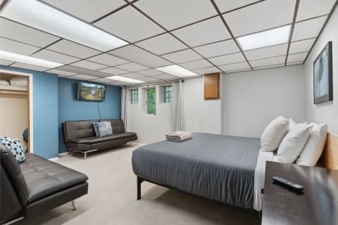 A modern basement bedroom featuring a gray bed, a sofa, and a TV mounted on the blue wall.