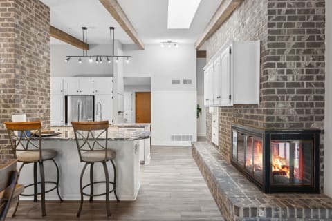 A modern kitchen with a granite island, brick fireplace, and wooden beams.