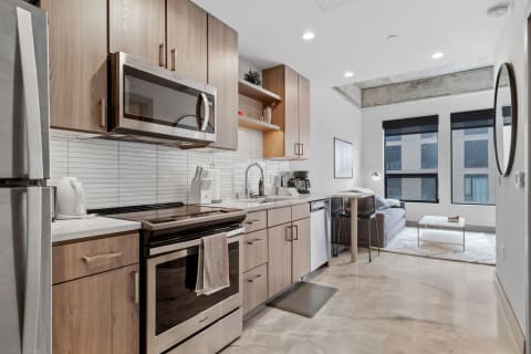 Contemporary kitchen with light wood cabinets and stainless steel appliances, featuring a cozy living area in the background.