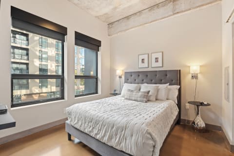 A modern bedroom featuring a gray bed with plush bedding, large windows, and urban views.