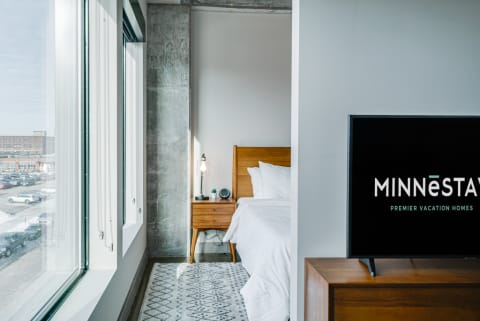 Interior view of a stylish bedroom with a large window, wooden furniture, and a mounted TV displaying a vacation rental logo.