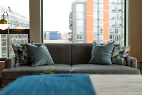 A gray couch with pillows in a room featuring a large window displaying an urban view.
