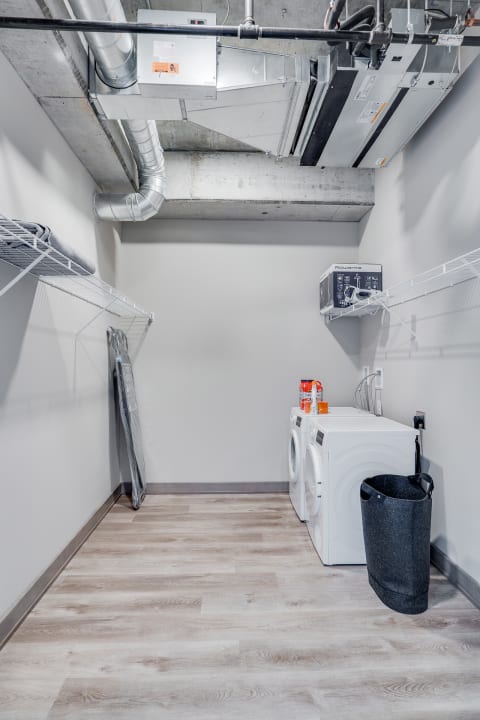A clean laundry room with white washing machines, a dark laundry basket, and a wire shelf.