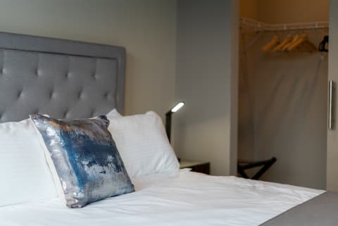A cozy hotel room with a tufted gray headboard, white bedding, and a decorative blue pillow, with a small desk lamp and closet visible.
