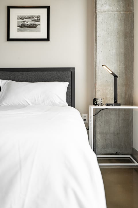 A modern bedroom with a gray upholstered headboard, white bedding, and a contemporary nightstand with a lamp and digital clock.