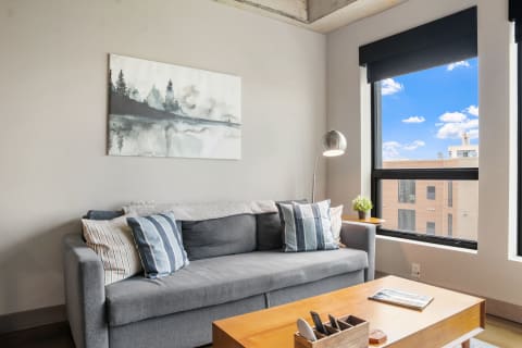 Cozy living room with a sofa, painting, and window showing a blue sky with clouds.