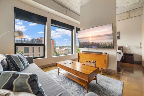 Modern living room with gray couch, wooden coffee table, and view of the city through large windows.