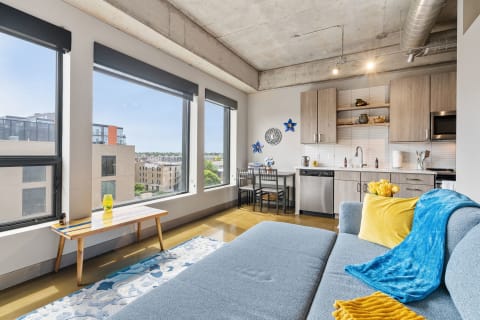 Modern apartment living area with large windows, a blue couch, and a kitchenette.