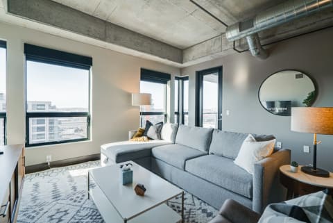A stylish living room featuring a grey sectional sofa, large windows, and an industrial concrete ceiling.