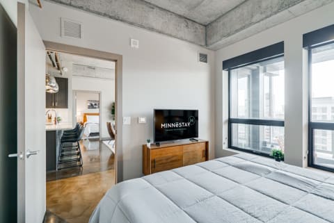 Contemporary bedroom with a bed, TV, and views of the city through large windows.
