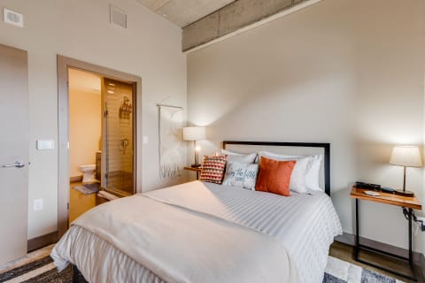 A well-decorated bedroom with a neatly made bed, bedside lamp, and an open bathroom door in view.