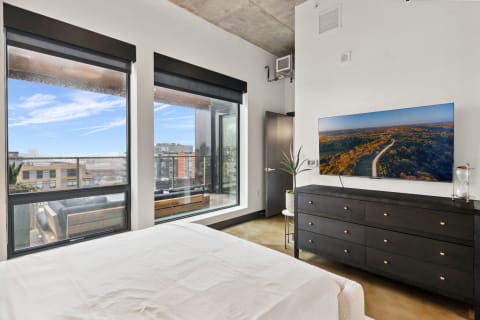 A contemporary bedroom featuring large windows, a dark dresser with a TV, and stylish decor.