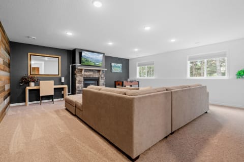 A modern living room with a large sectional sofa, a stone fireplace, and large windows overlooking nature.