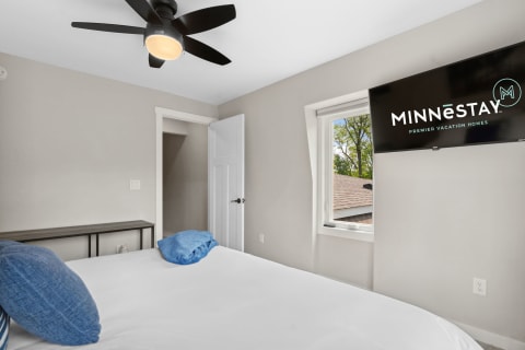 Cozy bedroom with white bed and blue pillows, flat-screen TV on wall displaying MINNēSTAY logo.