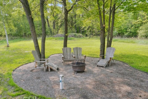 Outdoor seating area with four Adirondack chairs around a fire pit in a grassy landscape.