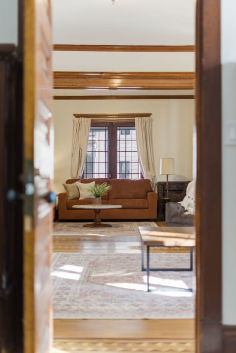 Living room viewed through an open door, featuring a couch, coffee table, and windows with curtains.