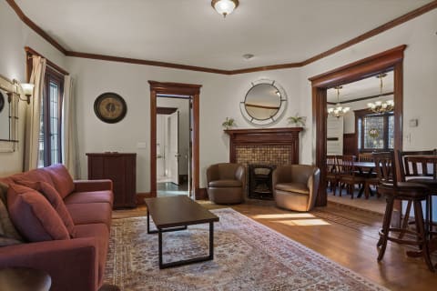 A comfortable living room with a burgundy sofa, leather armchairs, and a fireplace, leading to a dining area with a stained glass window.
