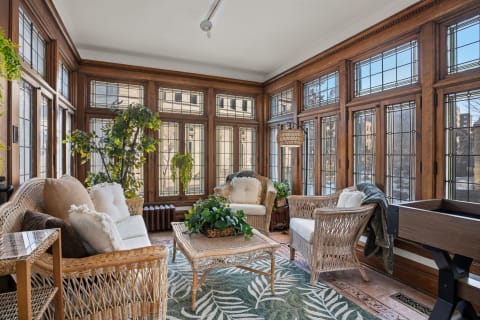 A cozy three-season conservatory with wicker furniture, lush plants, and large windows offering natural light.