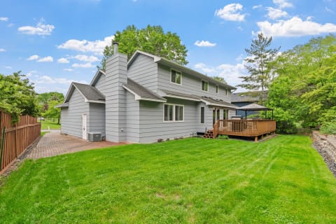 Two-story gray house with a wooden deck and green lawn under a blue sky.