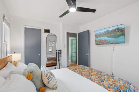 A bright bedroom featuring a bed with colorful bedding, a round mirror, and a television displaying a landscape.