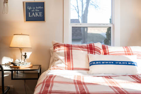 A cozy bedroom with plaid comforter, lake-themed sign, and bedside lamp.
