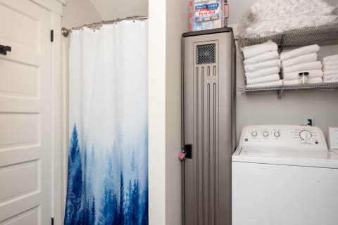 Laundry area featuring a blue tree-patterned shower curtain, a storage cabinet, and a washing machine, with stacked towels on a shelf.