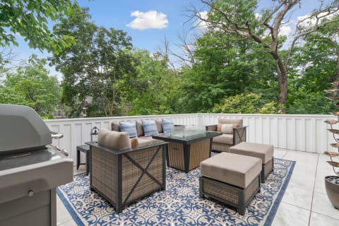 Outdoor patio featuring a cozy seating area, coffee table, and grill amidst green trees.