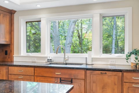 Contemporary kitchen featuring a sink, dark stone countertop, and large windows revealing trees outside.