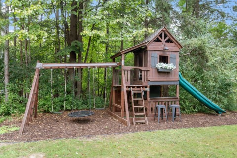 Wooden treehouse and swings surrounded by trees in a backyard.