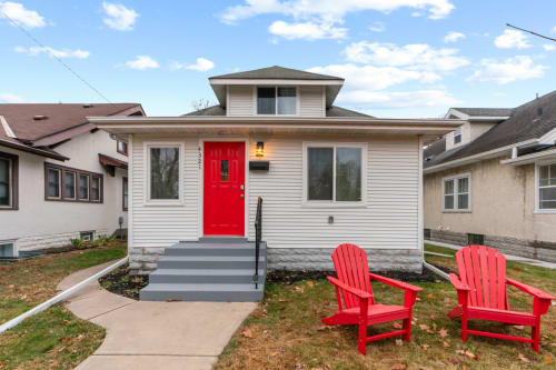 Charming home with vibrant red door and matching Adirondack chairs creating a welcoming arrival experience in a peaceful residential neighborhood.