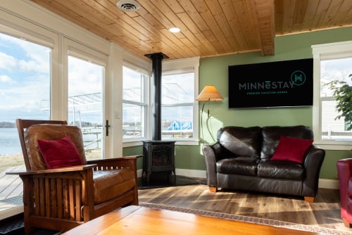 Cozy lakefront living room featuring leather seating, wood-burning fireplace, and stunning waterfront views through panoramic windows.